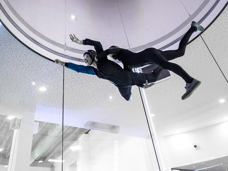Deux personnes en combinaison noire et casque dans un simulateur de chute libre, suspendues en l'air à l'intérieur d'une structure.