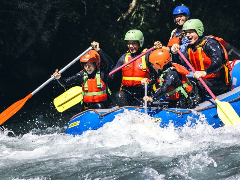Groupe de six personnes en rafting sur une rivière agitée, portant des casques verts et orange, pagayant avec détermination à travers des eaux tumultueuses, entourés d'éclaboussures d'eau blanche