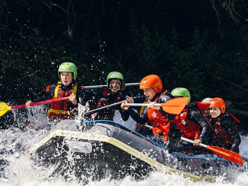 Famille de six personnes en rafting sur une rivière avec des rapides modérés, portant des casques de différentes couleurs et des gilets de sauvetage, pagayant ensemble avec des pagaies jaunes et roses