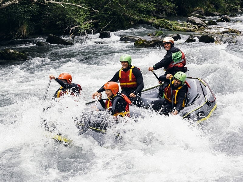 Groupe de six personnes en rafting sur une rivière agitée, portant des casques orange et verts, pagayant énergiquement parmi les rapides, entourés d'éclaboussures d'eau