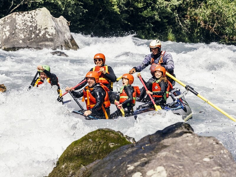 Équipe de six personnes en rafting descendant une rivière tumultueuse, portant des gilets de sauvetage orange et des casques, guidés par un instructeur, traversant des eaux vives avec des rochers