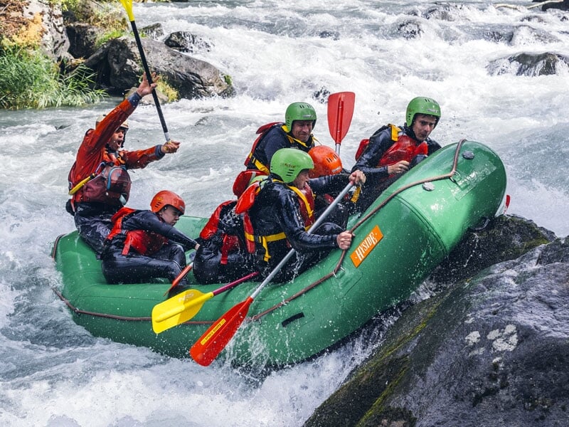 Six personnes en rafting sur une rivière rapide, utilisant des pagaies rouges et jaunes, portant des casques et des gilets de sauvetage, naviguant autour de gros rochers, l'eau bouillonnant autour d'e
