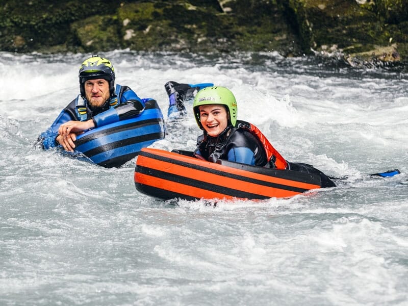 Deux personnes en hydrospeed descendant une rivière, portant des casques et des flotteurs rayés, l'une souriante tandis qu'elles naviguent les eaux agitées