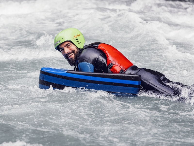 Homme en hydrospeed sur une rivière, portant un casque vert et un gilet de sauvetage rouge, souriant et tenant un flotteur rayé bleu et noir