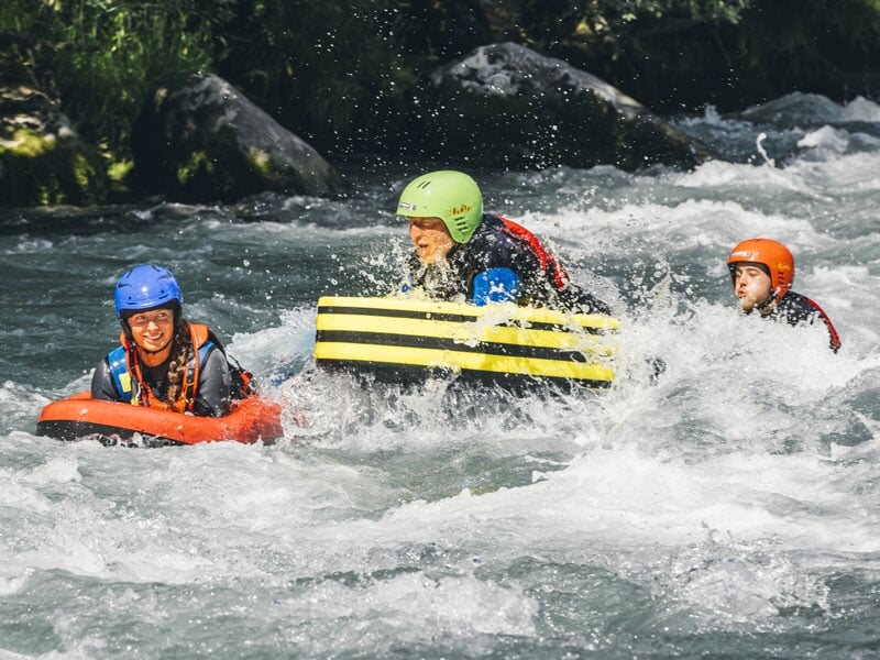 Trois personnes en hydrospeed descendant une rivière rapide, portant des casques colorés et tenant des flotteurs, éclaboussés par l'eau