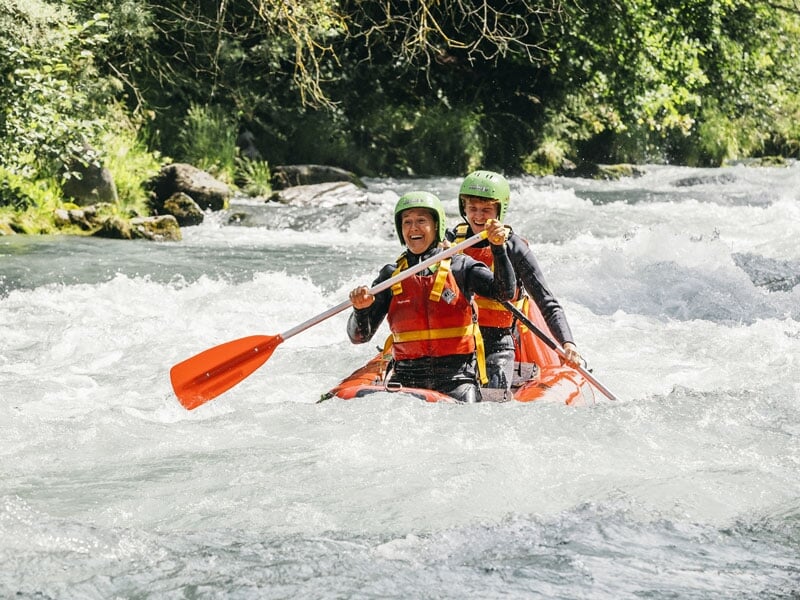 Deux personnes en canoë-kayak descendant une rivière avec des rapides modérés, portant des casques verts et pagayant avec des pagaies orange, souriantes et concentrées sur leur parcours