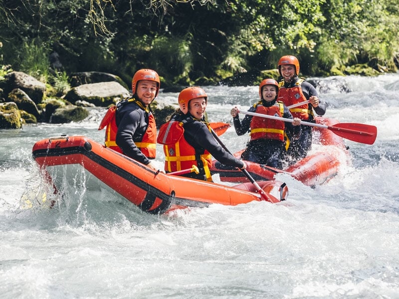 Quatre personnes en gilets de sauvetage et casques naviguent en raft sur une rivière agitée entourée de verdure dense.