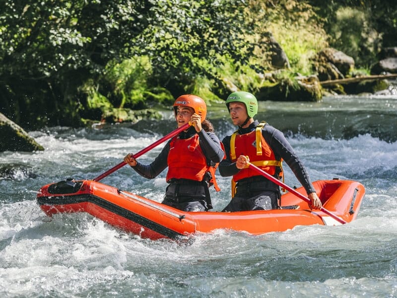 Deux personnes en canoë-kayak sur une rivière, portant des casques et des gilets de sauvetage, pagayant ensemble avec détermination, naviguant parmi les rapides et entourées de végétation dense