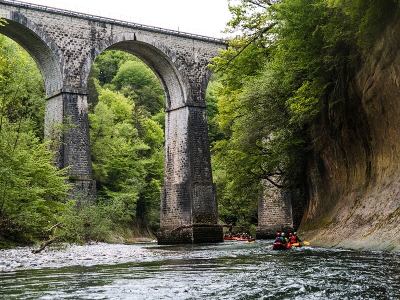 Vue d'un viaduc en pierre traversant une rivière, des canoës-kayaks avec des personnes portant des casques et des gilets de sauvetage pagayant en dessous, entourés de falaises et de verdure