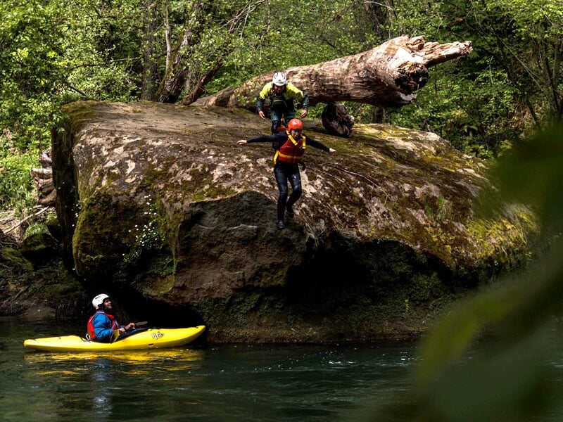 Personne sautant d'un rocher dans une rivière tandis qu'une autre attend son tour, portant des combinaisons et des casques, un kayak jaune avec une personne à l'intérieur les attendant dans l'eau, ent