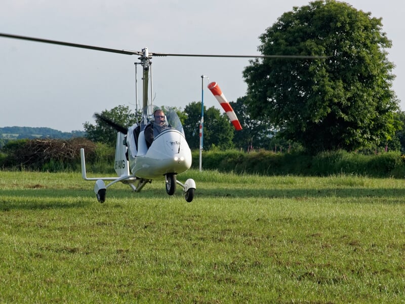 Un ULM blanc posé sur un champ vert avec un pilote souriant, un arbre et un mât avec une manche à air en arrière-plan.