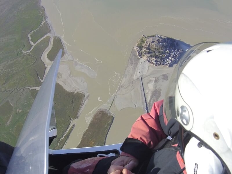Vue aérienne prise depuis un ULM montrant un pilote avec casque blanc et un paysage côtier avec un rocher et de l'eau.
