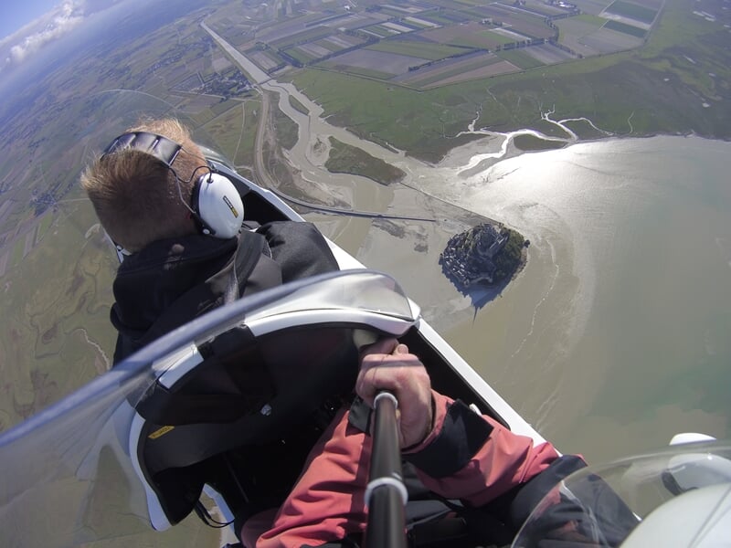 Pilote en ULM avec casque et casque audio, tenant un selfie stick, survolant un paysage côtier avec un rocher et de l'eau.