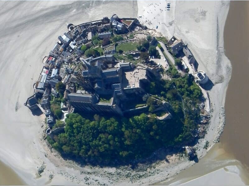 Vue aérienne rapprochée d'une île fortifiée entourée de sable et d'eau, prise depuis un ULM en vol au-dessus de la côte.