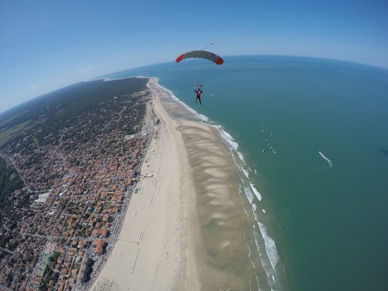 Parachutiste en chute libre au-dessus d'une longue plage bordée par une ville et l'océan sous un ciel bleu.