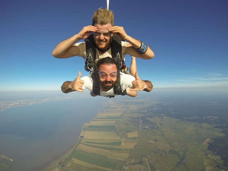 Deux personnes en tandem en parachutisme souriantes en chute libre au-dessus d'un paysage rural et d'un plan d'eau.
