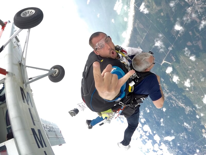 Saut en parachute en tandem avec un homme faisant un geste de la main en signe de rock, ciel et nuages en arrière-plan.