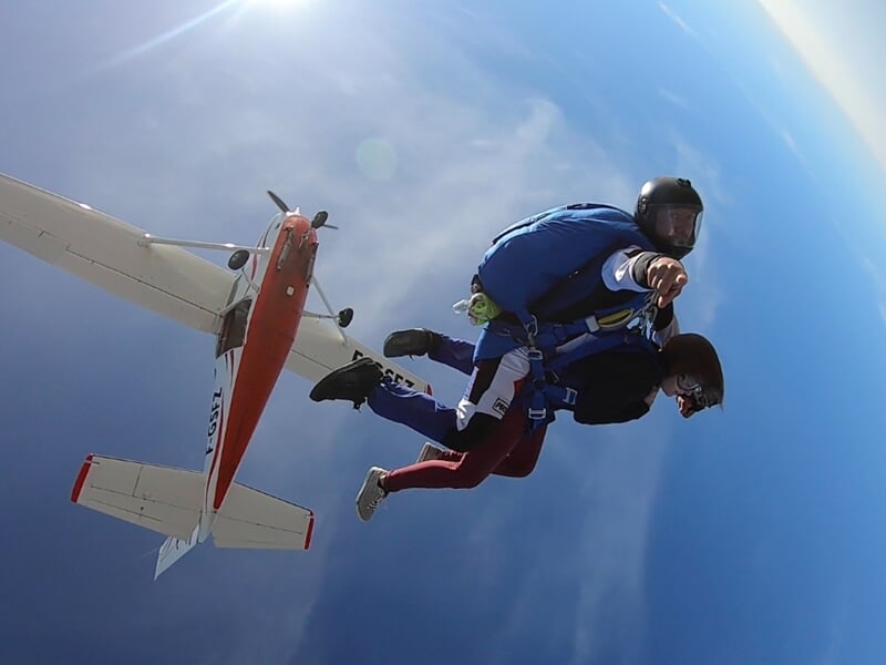 Deux parachutistes en tandem en chute libre près d'un avion rouge et blanc avec un ciel bleu clair en arrière-plan.