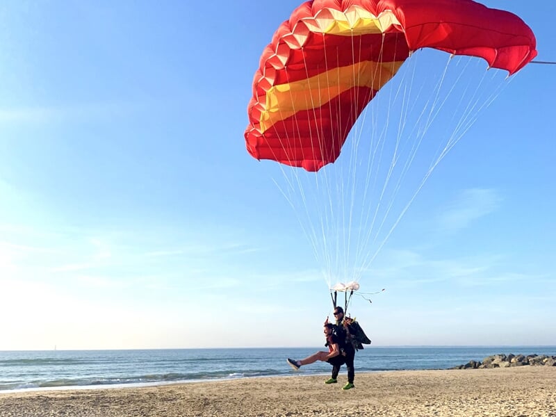 Parachutiste atterrissant sur une plage avec un parachute rouge et jaune, mer calme et ciel bleu en arrière-plan.