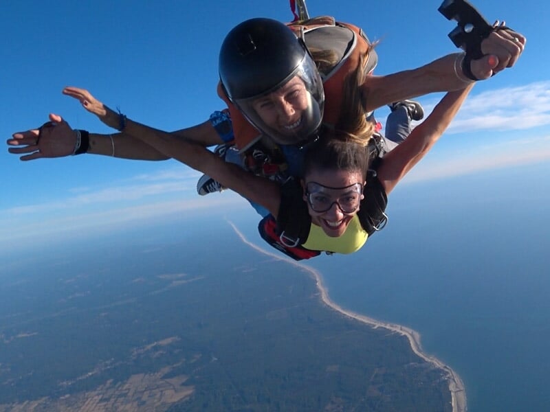 Parachutistes en tandem souriants en chute libre au-dessus d'une côte avec une plage et la mer sous un ciel clair.