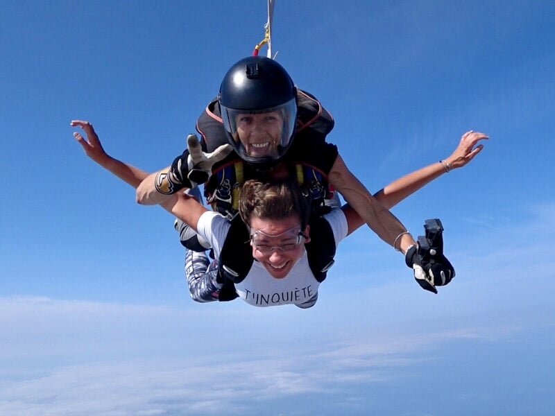 Deux parachutistes en tandem souriants en chute libre avec un paysage côtier visible au loin sous un ciel bleu.