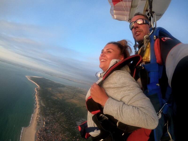 Parachutistes en tandem en chute libre au-dessus d'une plage et d'une mer avec un ciel bleu et quelques nuages.