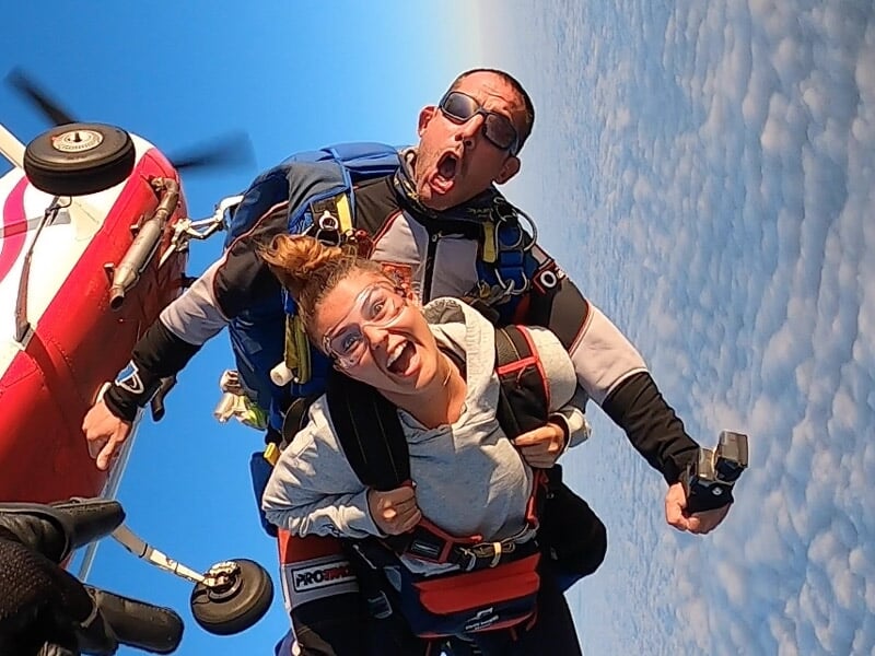 Femme en parachutisme en tandem souriante avec un instructeur, ciel bleu et nuages en arrière-plan.