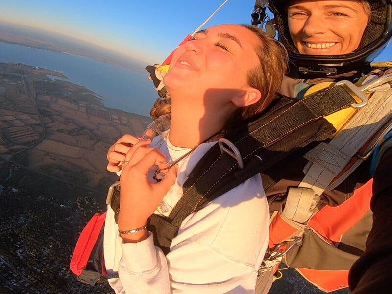 Femme en parachutisme en tandem souriante avec un instructeur, ciel bleu et nuages en arrière-plan.