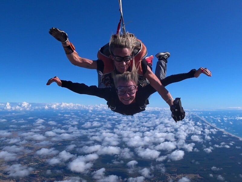 Deux personnes en parachutisme en tandem en chute libre au-dessus d'un paysage avec des nuages et un ciel bleu.