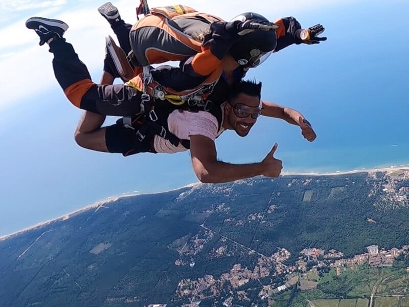 Parachutiste en tandem faisant un signe de pouce levé, avec une vue panoramique sur des forêts et des zones urbaines en bord de mer.