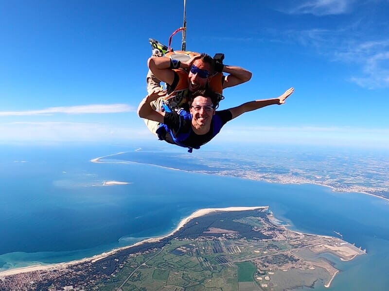 Deux parachutistes en tandem souriants, survolant une côte bordée de plages, d'océan bleu et de paysages verdoyants.