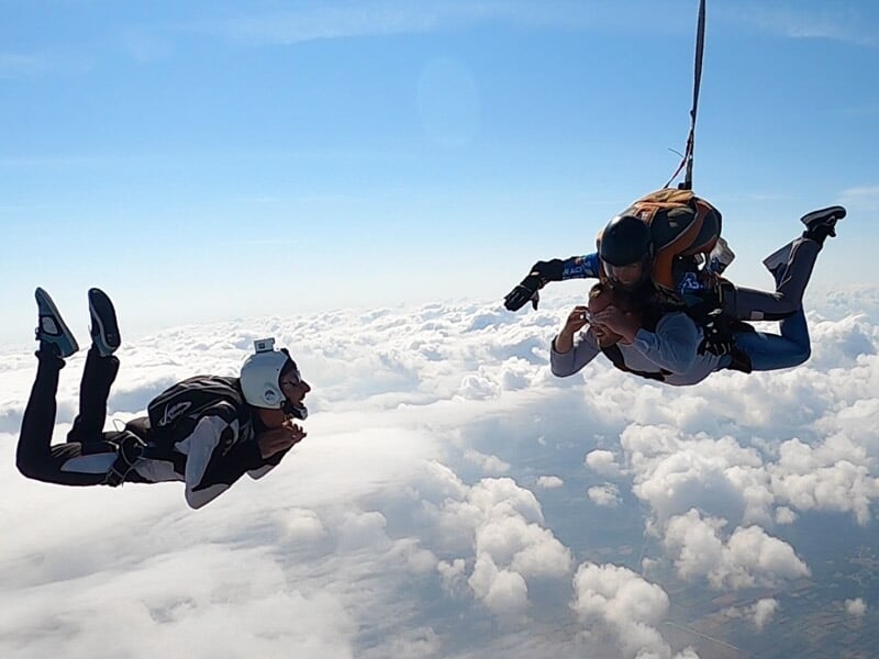 Tandem de parachutistes en chute libre, entourés de nuages blancs, avec un accompagnateur à proximité dans un ciel dégagé.