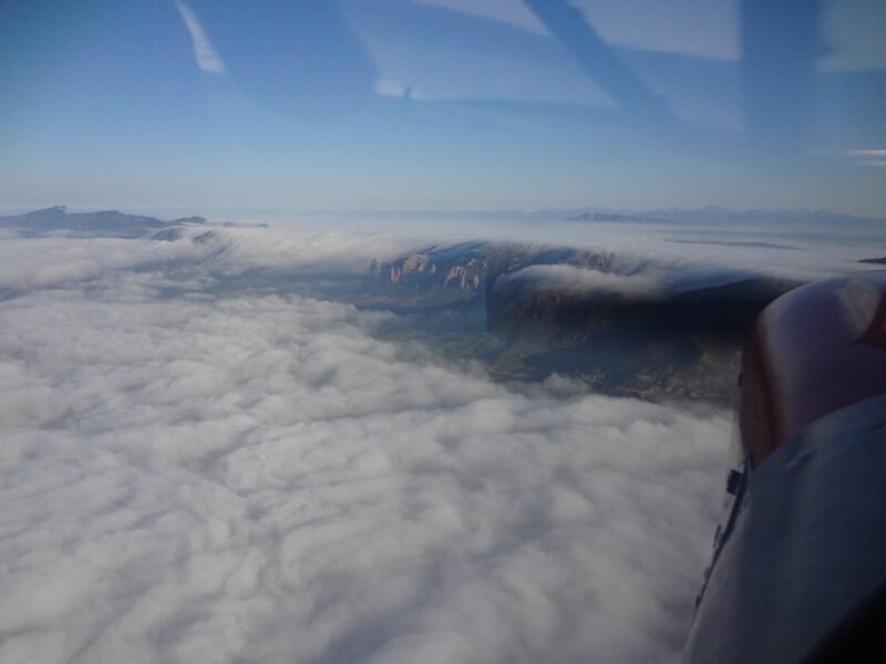 Vue aérienne au-dessus des nuages avec un paysage montagneux partiellement visible sous un ciel bleu clair.