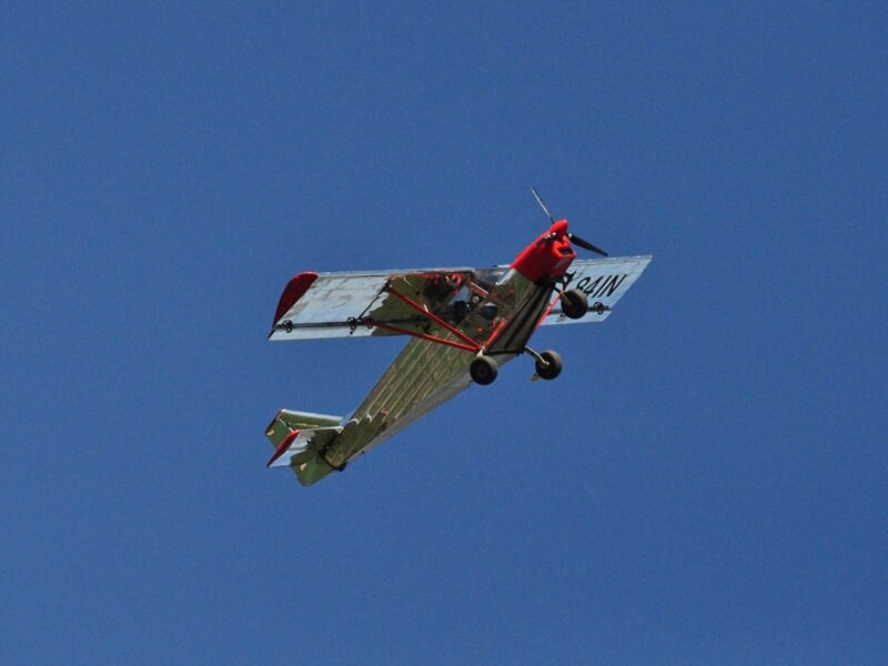 Petit avion ultraléger rouge et blanc en vol dans un ciel bleu dégagé, vu de dessous et de côté.