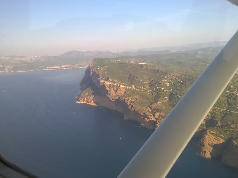 Paysage côtier vu depuis un avion, avec falaises et mer calme sous un ciel partiellement nuageux.