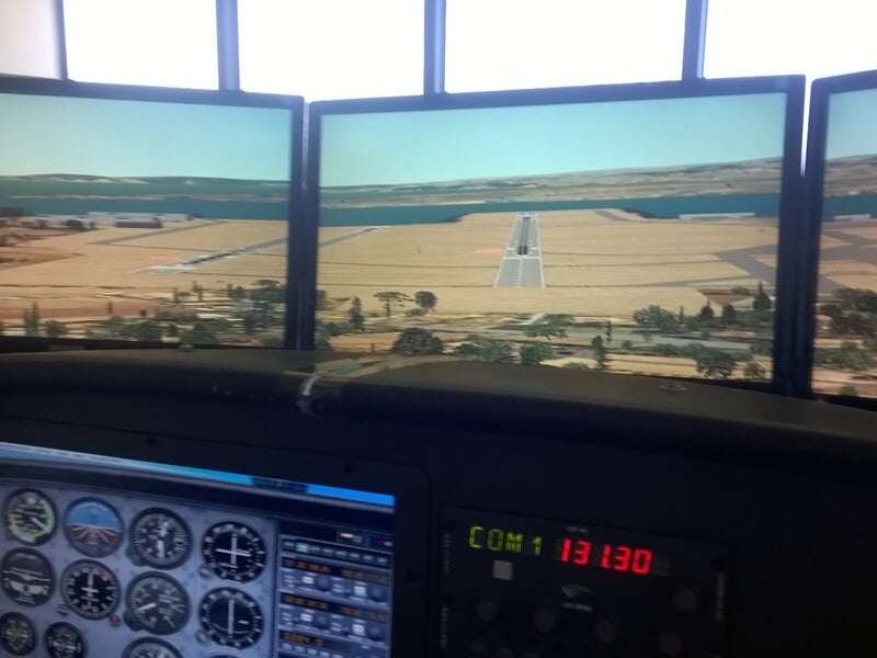 Cockpit d'un simulateur de vol avec plusieurs écrans affichant une piste d'atterrissage et des commandes.