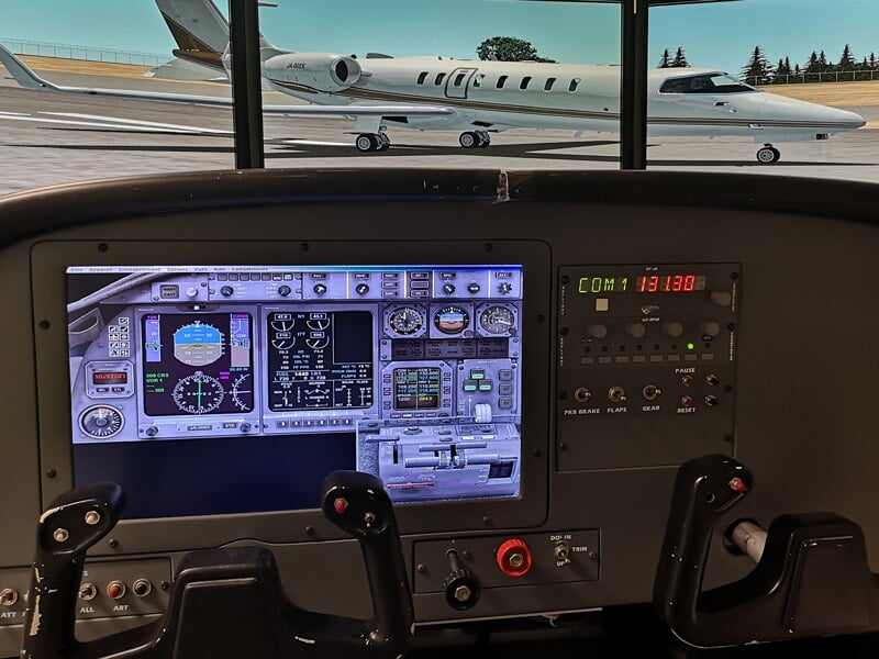 Cockpit d'un simulateur de pilotage avec écrans et commandes, vue sur un jet privé à l'arrêt.
