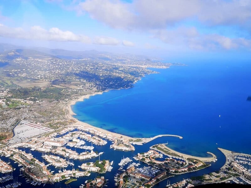 Vue aérienne d'une côte méditerranéenne avec plages, port et collines sous un ciel bleu clair.