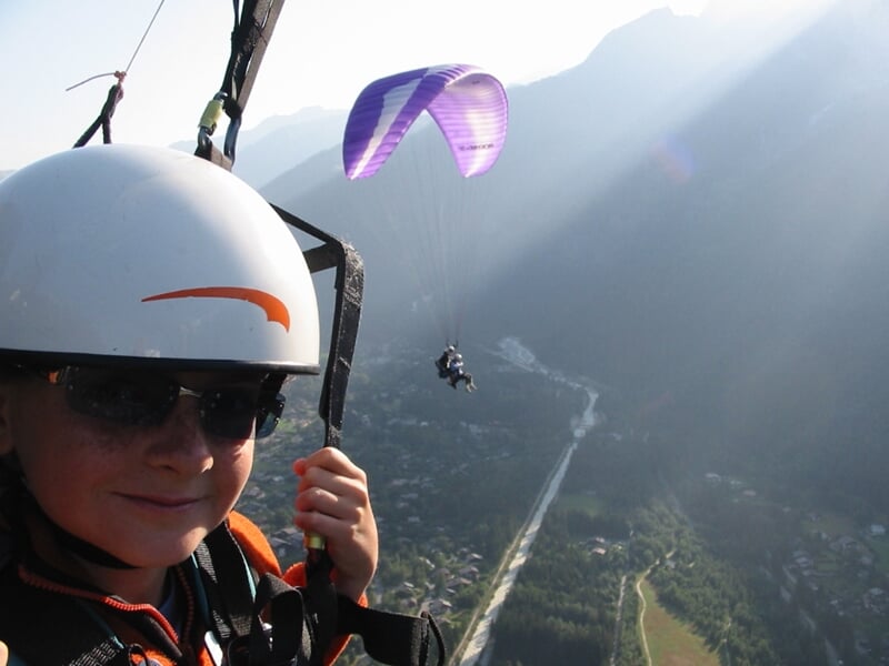 Jeune garçon en casque et lunettes de soleil en parapente au-dessus d'une vallée montagneuse ensoleillée.