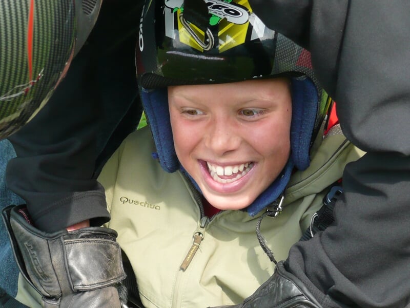 Enfant souriant portant un casque de protection, prêt pour un baptême de parapente encadré par un adulte.