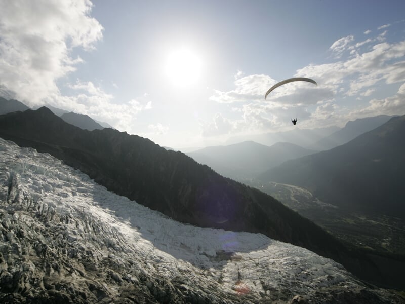 Parapente volant au-dessus d'un glacier et des montagnes sous un ciel partiellement nuageux et lumineux.