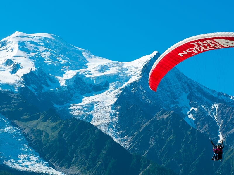 Parapente rouge volant devant le Mont Blanc avec un ciel bleu clair et un paysage montagneux spectaculaire.