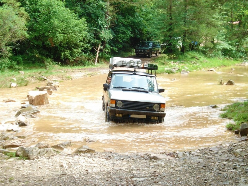 Véhicule tout-terrain blanc traversant une rivière peu profonde dans un environnement naturel boisé.