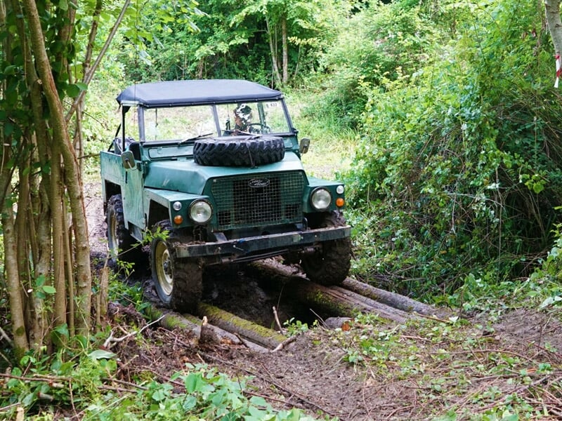 Véhicule tout-terrain vert foncé traversant un chemin boueux en forêt avec des troncs d'arbres.