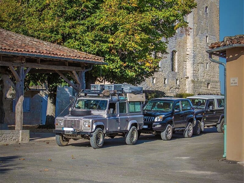 Véhicule tout-terrain gris garé dans un village avec des maisons anciennes et un clocher en arrière-plan.