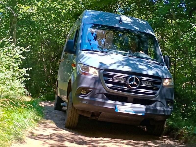 Fourgon Mercedes gris roulant sur un chemin forestier étroit entouré d'arbres verts en plein jour