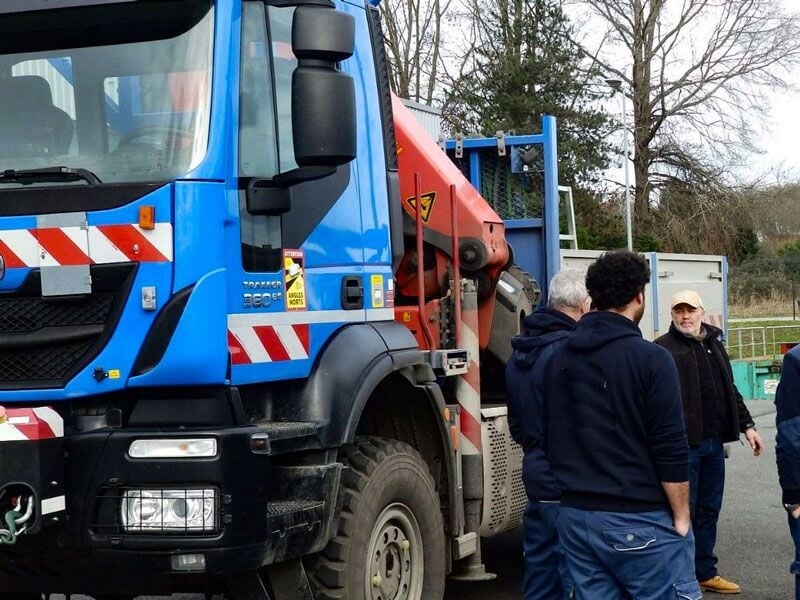 Gros plan sur un camion bleu avec une grue, plusieurs personnes discutent à côté du véhicule
