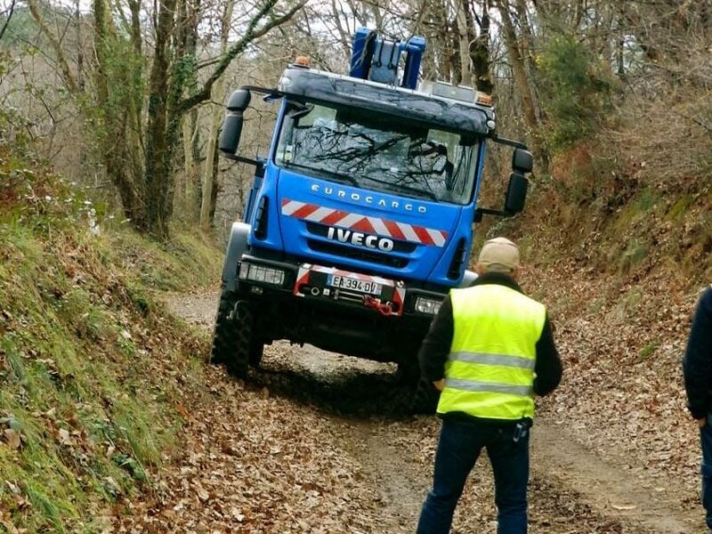 Camion bleu tout-terrain sur un chemin en forêt, un homme en gilet jaune observe la progression