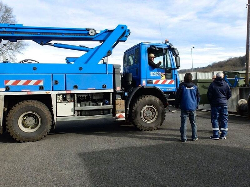 Camion bleu avec grue stationné sur une surface goudronnée, trois personnes debout à proximité