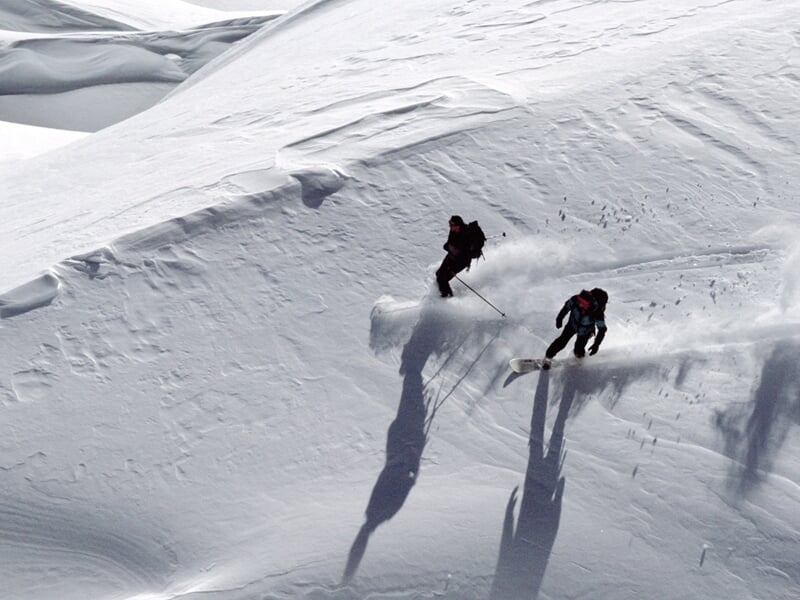 Deux skieurs dévalant une pente enneigée en montagne sous un ciel clair, laissant des traces dans la poudreuse.